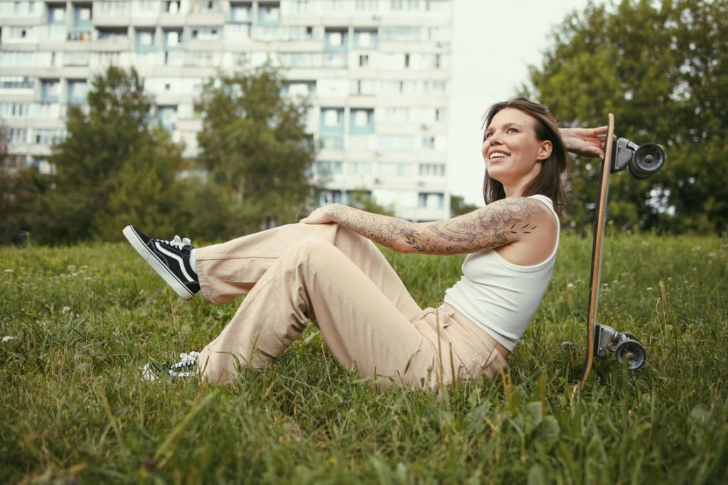 Девушка со скейтбордом за спиной сидит на траве